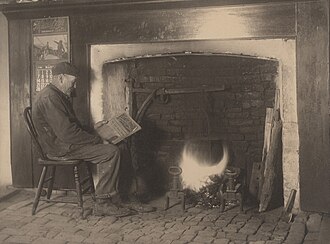 photographie représentant un homme lisant un journal près d'un feu de cheminée.